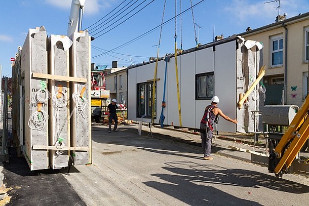 Serielle Sanierung von Energiesprong (drei Fassaden-Module auf der Straße abgestellt, daneben ein Modul am Kran mit Bauarbeitern vor einem Reihenhaus).