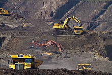 Gelbe Bagger graben in Kohle-Abbaugebiet. Rauch steigt von braunem Boden auf.